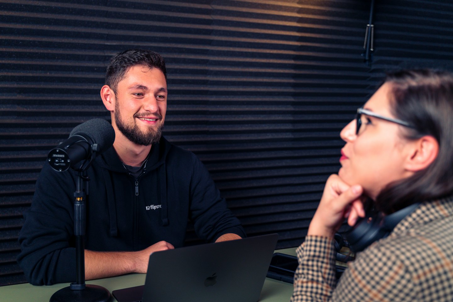Man speaks into microphone during podcast recording, woman listens nearby.